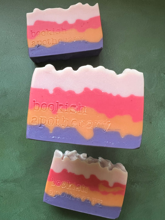 Three bars of soap with layers of pink, red, orange and purple stamped with bookish apothecary branding.