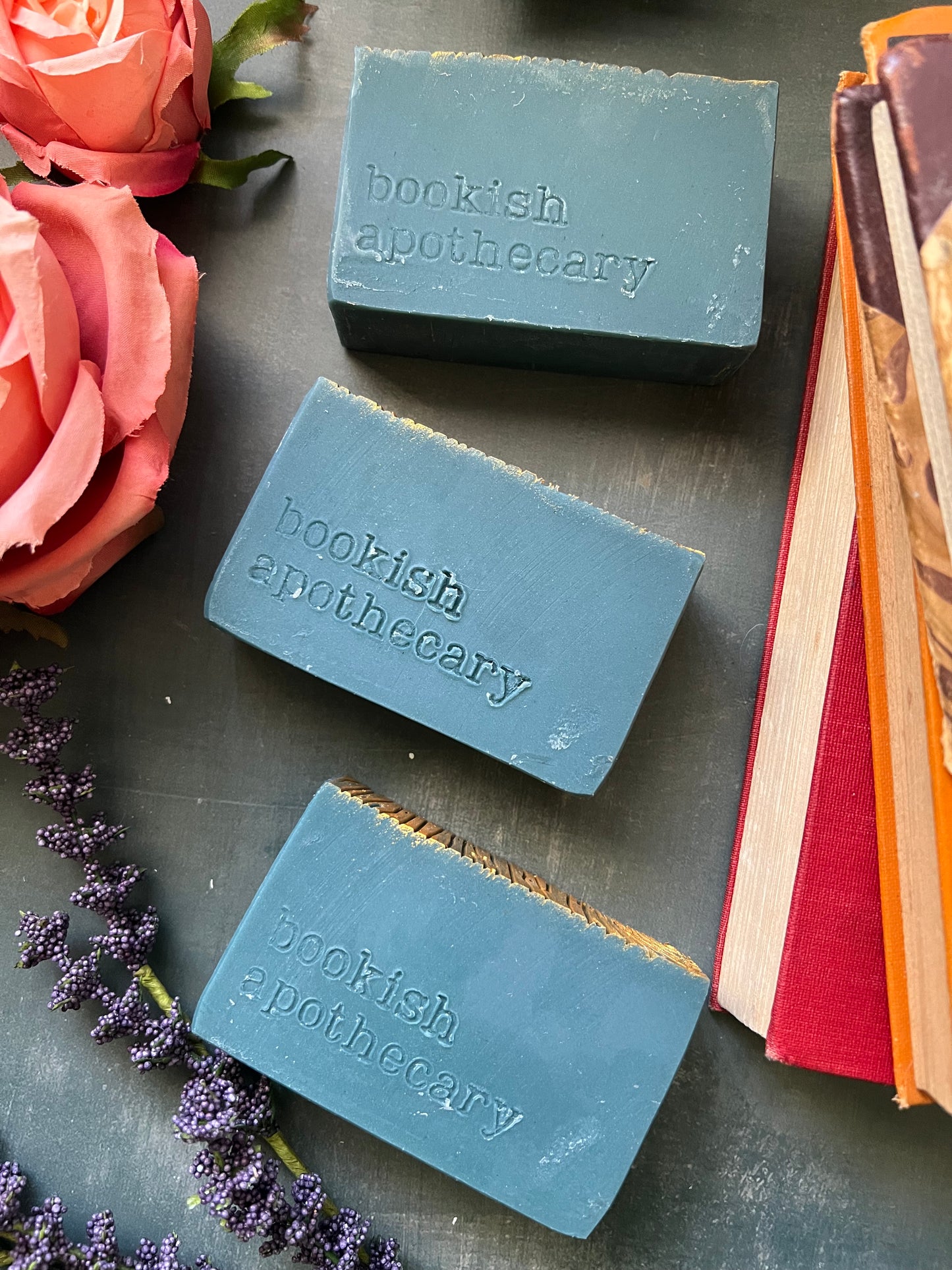 Three blue soap bars with 'bookish apothecary' branding on a dark surface with flowers and books.