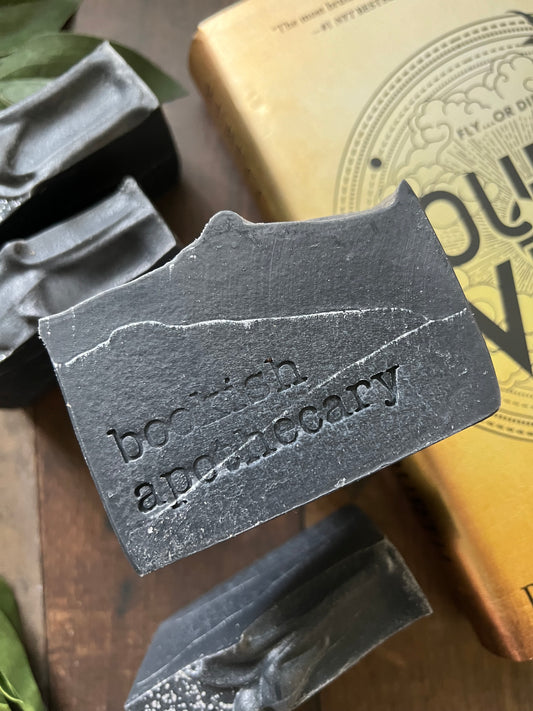 A bar of handmade soap with a gray color and speckled with dark dots, placed on a wooden surface with green leaves in the background and a label that reads 'COLLECTOR EDITION online exclusive'.
