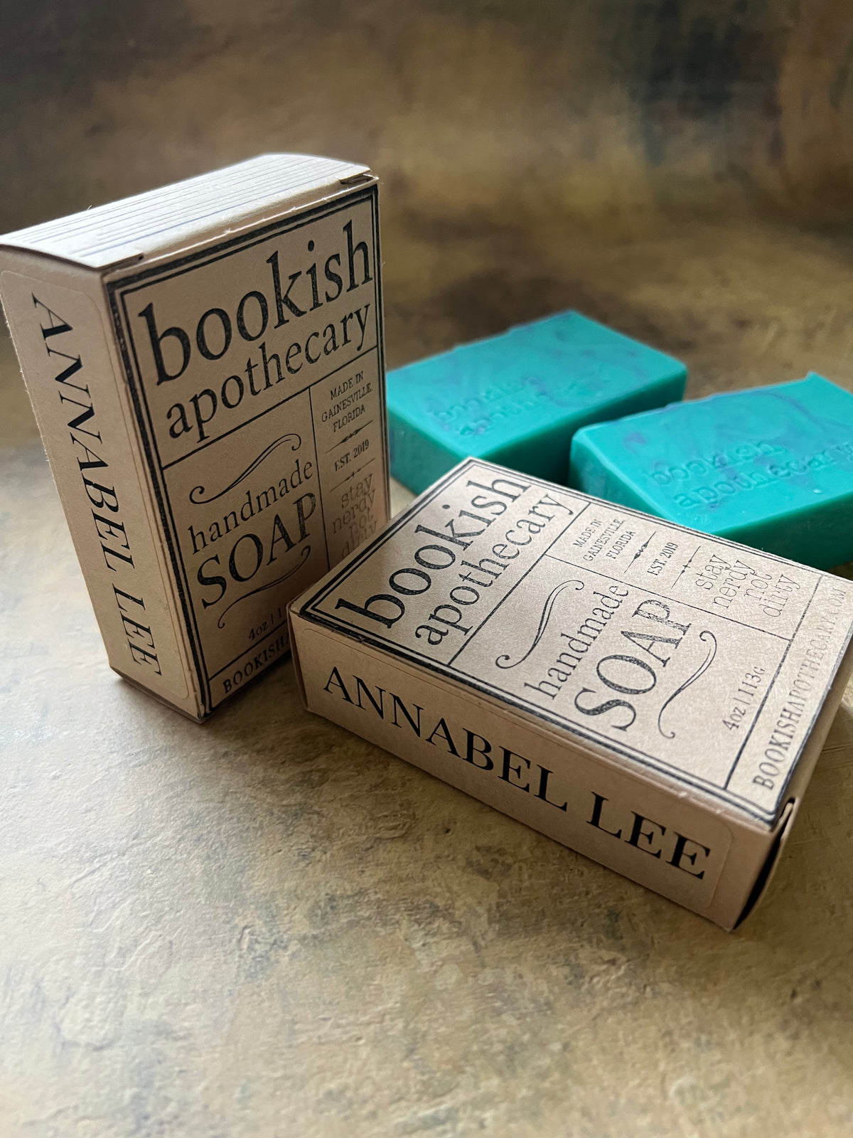 Two boxes of Annabel Lee Bookish Apothecary handmade soap displayed on a textured stone surface, with two turquoise-colored soap bars stacked behind them.