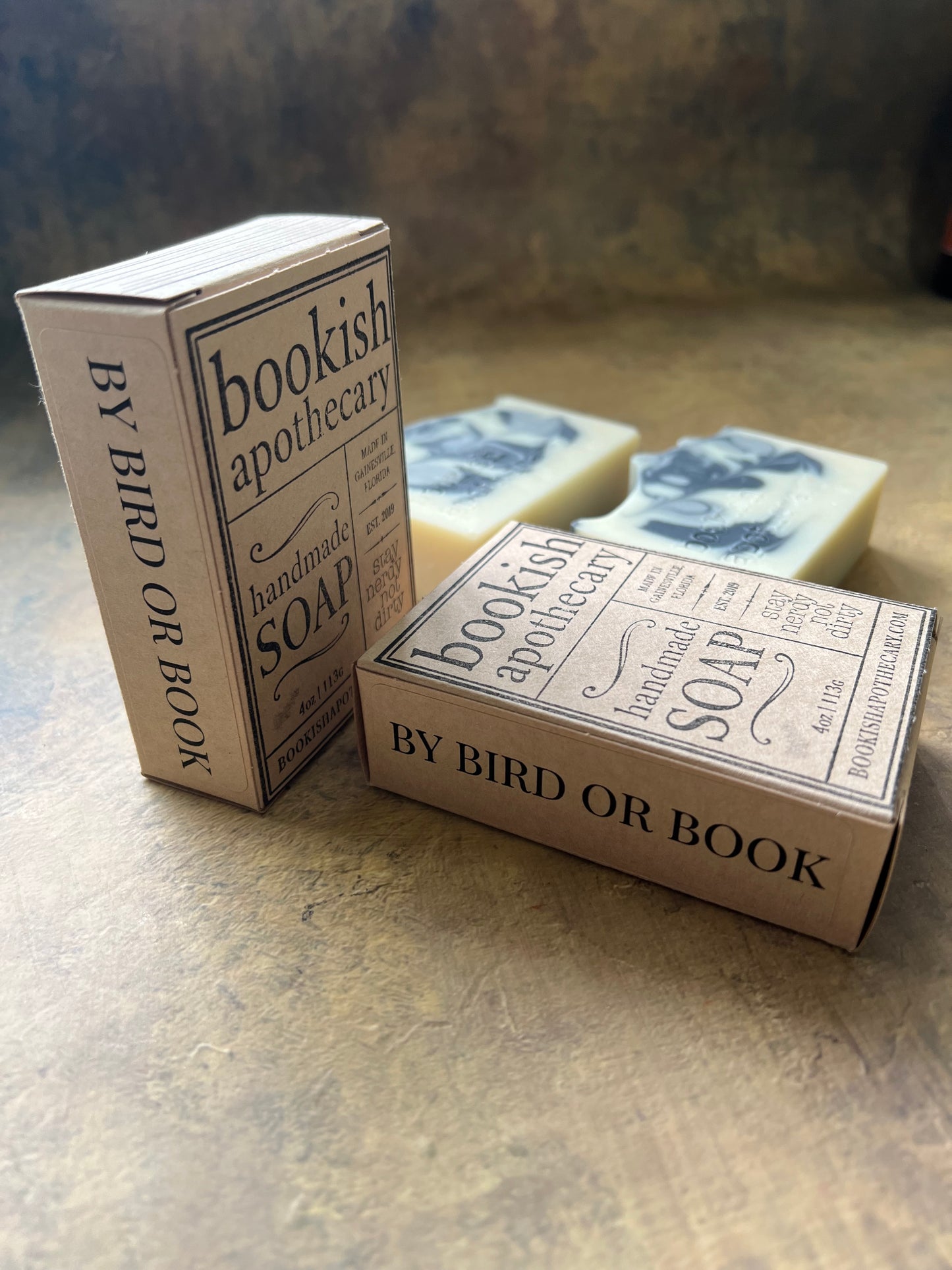 Two boxed bars of handmade soap labeled 'Bookish Apothecary' and 'BY BIRD OR BOOK' arranged on a textured beige surface.