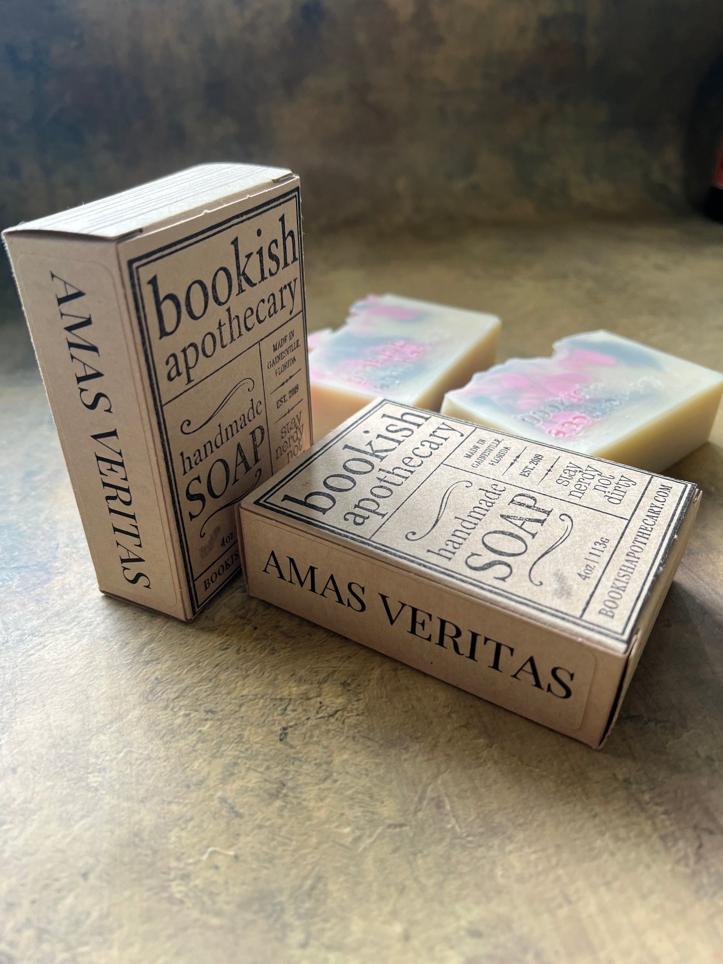 Two bars of handmade apothecary soap in decorative paper boxes with vintage-style labels, branded AMAS VERITAS and Bookish Apothecary, arranged on a textured stone surface.