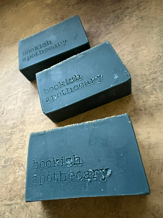 Three dark green rectangular soap bars with 'bookish apothecary' stamped on them, arranged on a textured beige surface.