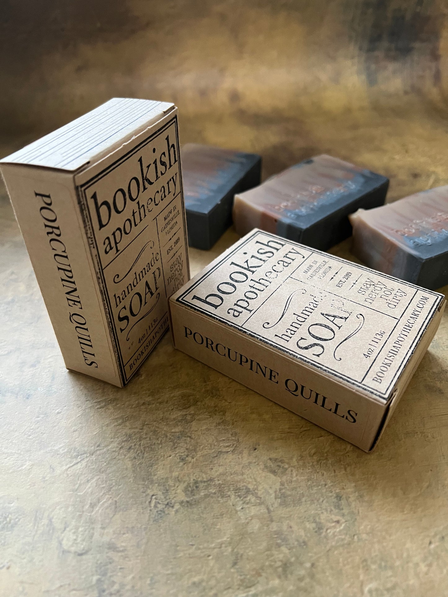 Two boxed bars of soap labeled 'Bookish Apothecary' arranged on a textured beige surface, with additional stacked boxes blurred in the background.