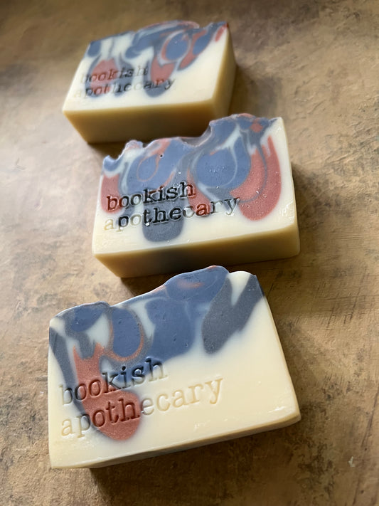 Three handmade artisanal soap bars with cream, blue, and red swirls, embossed with the text 'bookish apothecary', arranged diagonally on a textured brown surface.