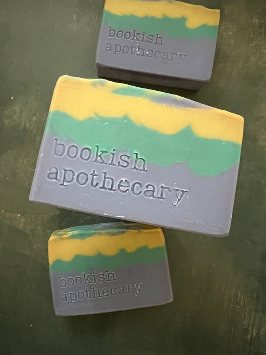 Three handmade soap bars with layers of yellow, green and blue featuring the brand name 'bookish apothecary' stamped on the front, displayed on a dark green surface.