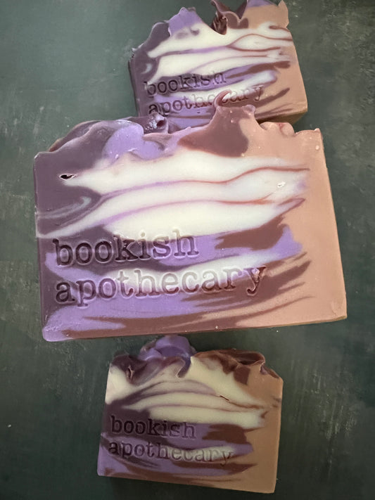 Three bars of handmade soap with purple, lavender, and white marbled swirls, stamped with the brand name 'bookish apothecary', displayed on a dark gray textured surface.