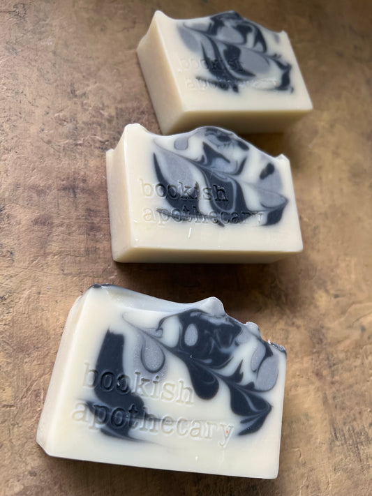 Three rectangular bars of soap with a marbled black and gray pattern on an off-white base, arranged diagonally on a textured brown surface. The bars are stamped with the brand name 'bookish apothecary'.