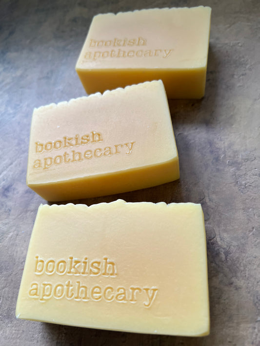 Three pale yellow rectangular bars of handmade soap with scalloped edges, stamped with the brand name 'bookish apothecary', arranged on a textured gray surface.