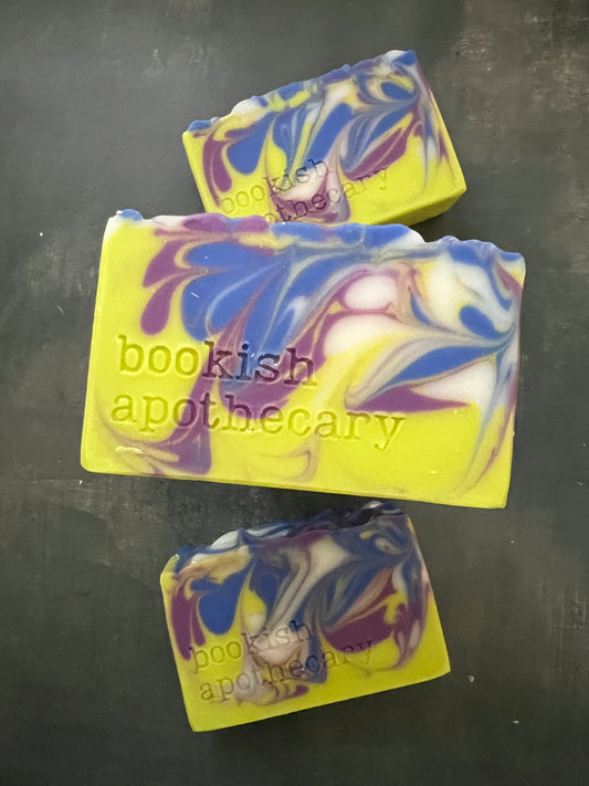 Three handmade soap bars with a marbled swirl pattern of chartreuse, blue, purple, and white, featuring the stamped text 'bookish apothecary' on a dark gray background.