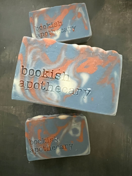 Three handmade soap bars with a blue-gray base and orange swirls, each stamped with the brand name 'bookish apothecary', arranged on a dark textured surface.