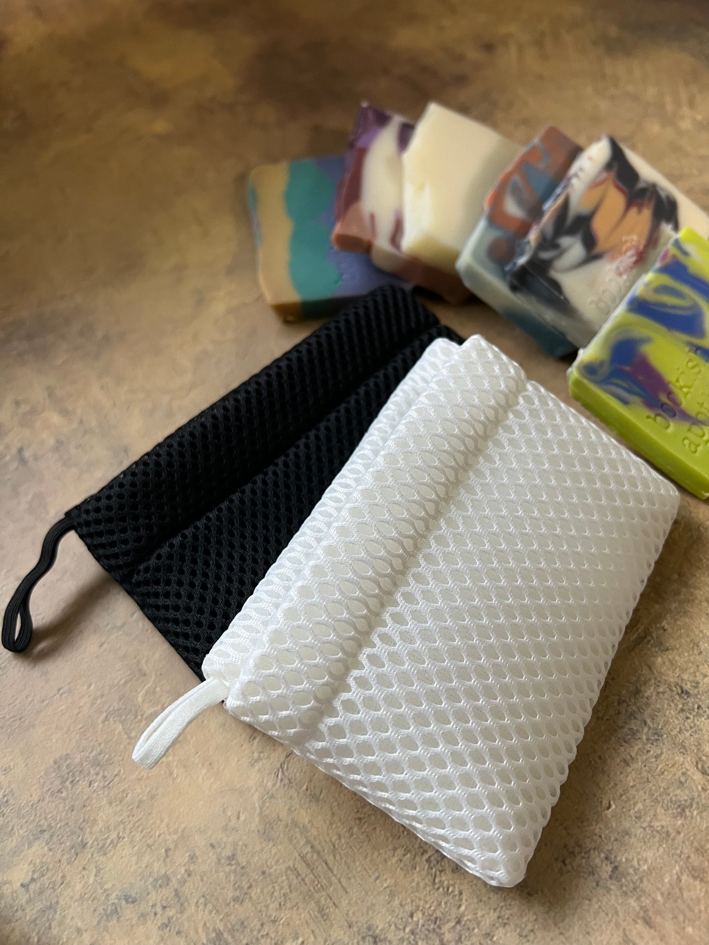 Two reusable wash or soap bags, one white with a raised honeycomb texture and one black with a mesh texture, shown with several colorful handmade soap bars on a textured beige surface.