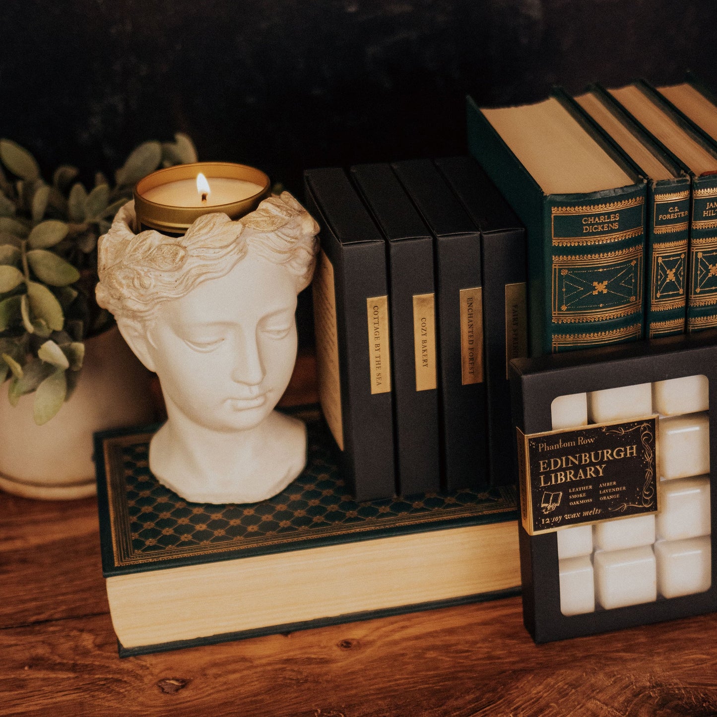 Decorative setup with a bust candle holder, books, and black boxes containing wax melts on a wooden surface.