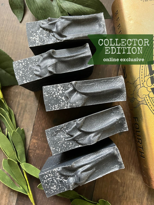 A bar of handmade soap with a gray color and speckled with dark dots, placed on a wooden surface with green leaves in the background and a label that reads 'COLLECTOR EDITION online exclusive'.