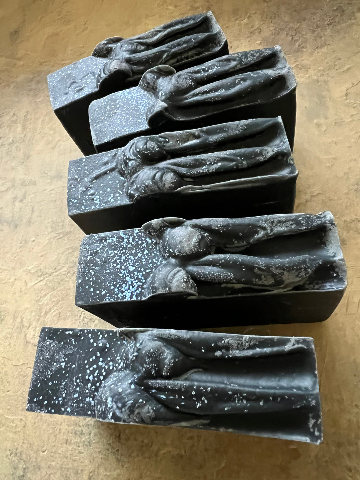 Five rectangular dark gray to black bars with raised organic sculptural tops, arranged on a textured tan surface.
