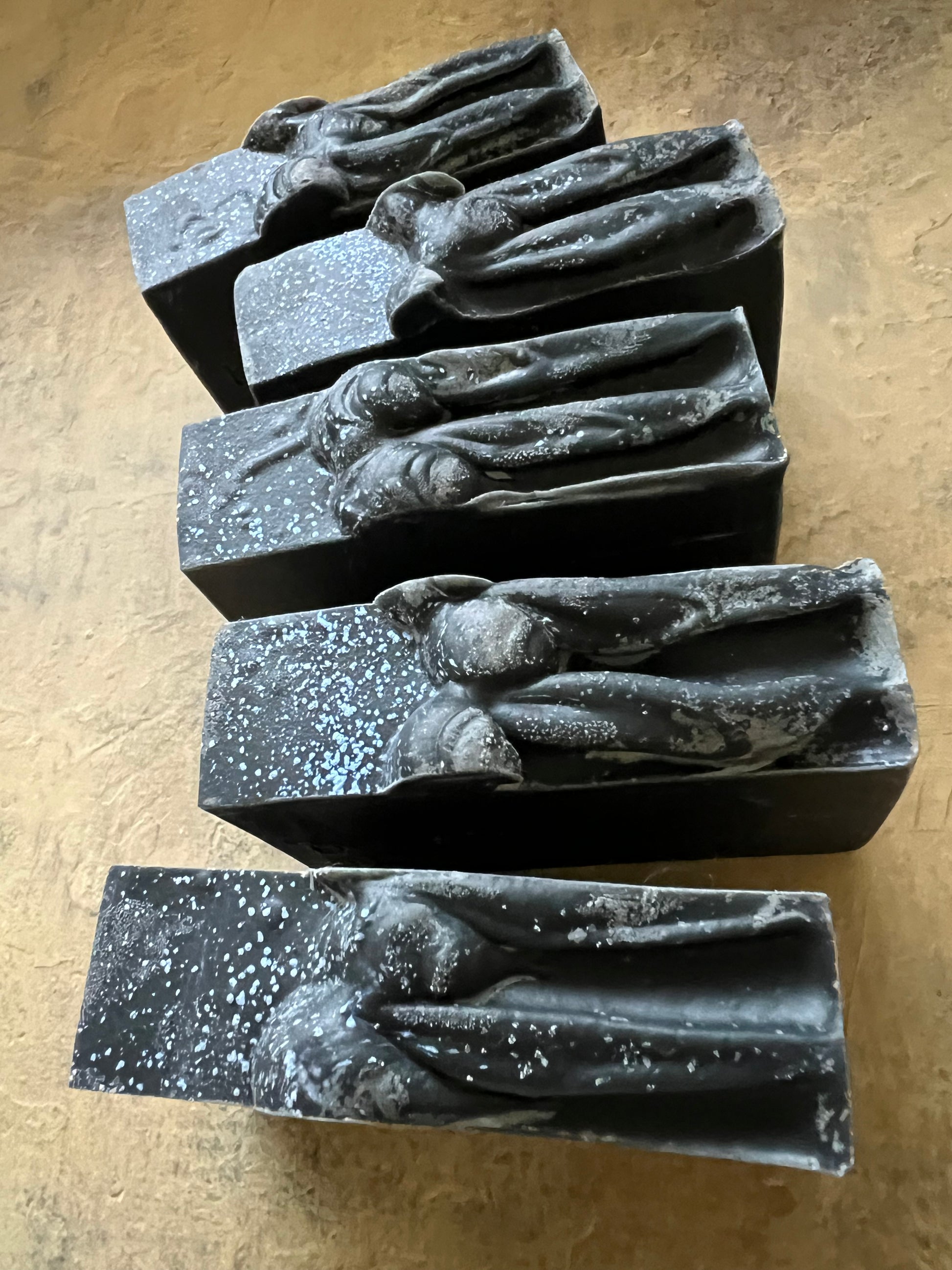 Five rectangular dark gray to black bars with raised organic sculptural tops, arranged on a textured tan surface.