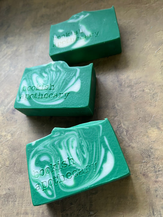 Three green handmade soap bars with white marbled swirls, stamped with the brand name 'bookish apothecary', displayed on a textured brown surface.