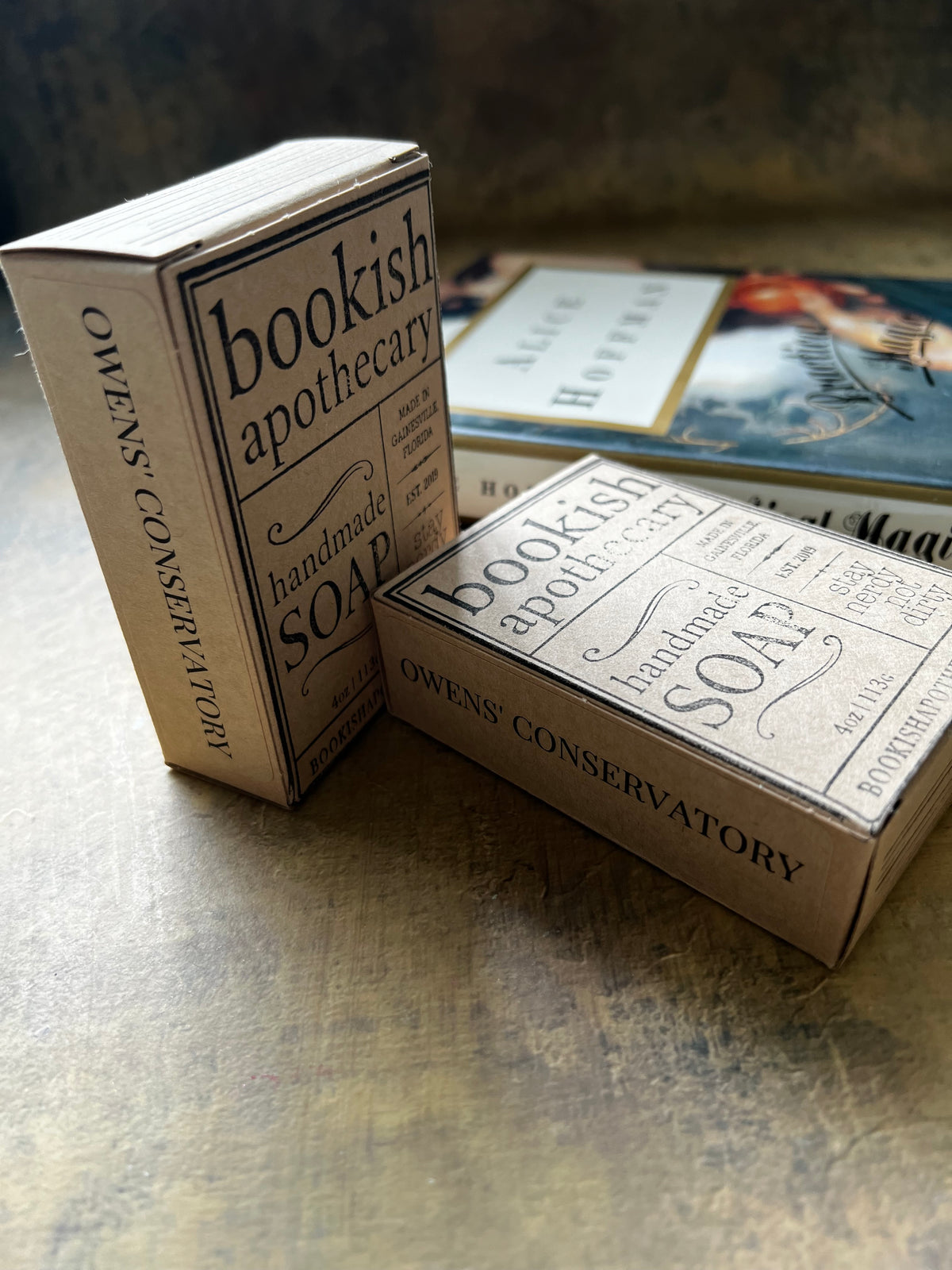 Two vintage-style soap boxes from Owens Conservatory labeled 'Bookish Apothecary Handmade Soap' arranged on a rustic wooden surface, with a blurred book in the background.