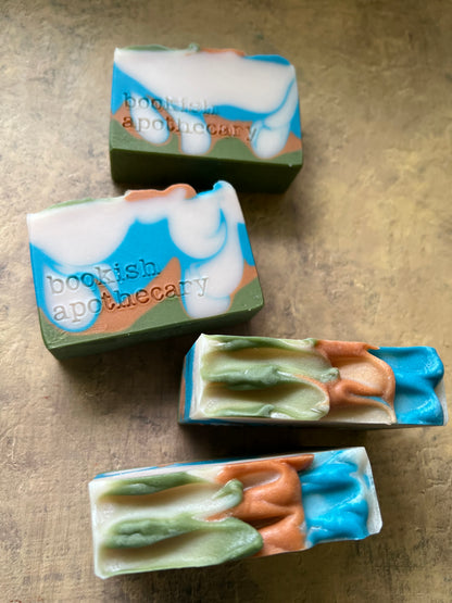 Four handmade marbled soaps with blue, green, peach, and white swirls, featuring the text 'bookish apothecary' stamped vertically, arranged on a textured beige surface.