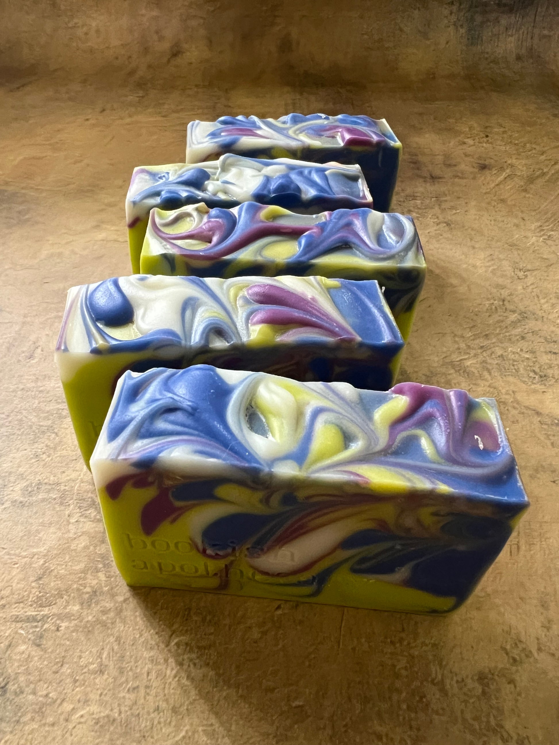 Four rectangular handmade soap bars with marbled swirls of blue, purple, white, and yellow, arranged diagonally on a textured brown surface.