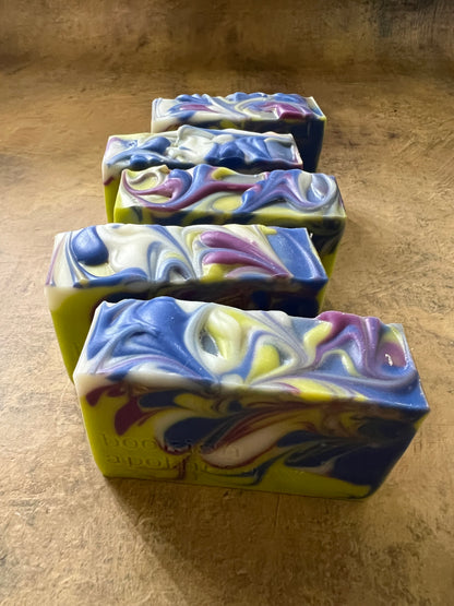 Four rectangular handmade soap bars with marbled swirls of blue, purple, white, and yellow, arranged diagonally on a textured brown surface.