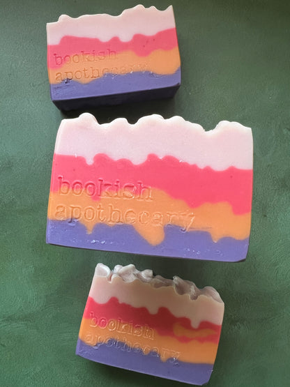 Three bars of soap with layers of pink, red, orange and purple stamped with bookish apothecary branding.
