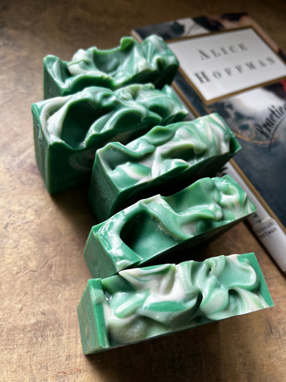 Five handmade green soap bars with white swirls are arranged diagonally on a textured brown surface next to a book.
