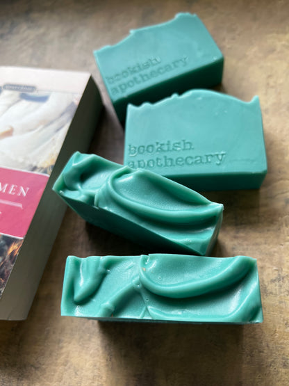 Four green handmade soap bars with wavy edges and the brand name 'bookish apothecary' embossed on them, arranged on a brown textured surface next to a book.