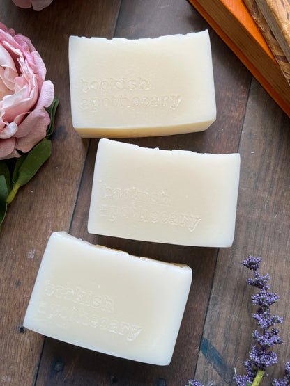 Three bars of soap on a wooden surface with flowers and a book in the background