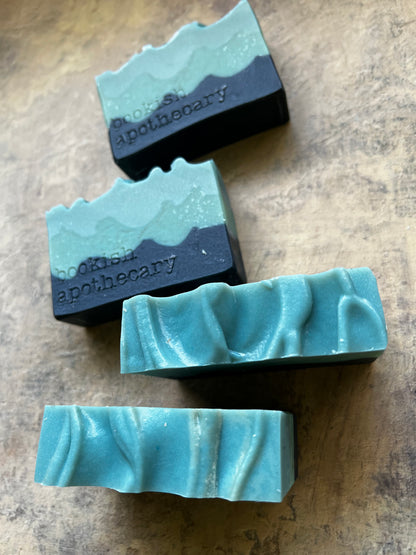Three handmade bar soaps with a two-tone wave design in turquoise and black, resting on a textured brown surface. The top soap bar is angled, showing the embossed brand name 'cookish apothecary'.