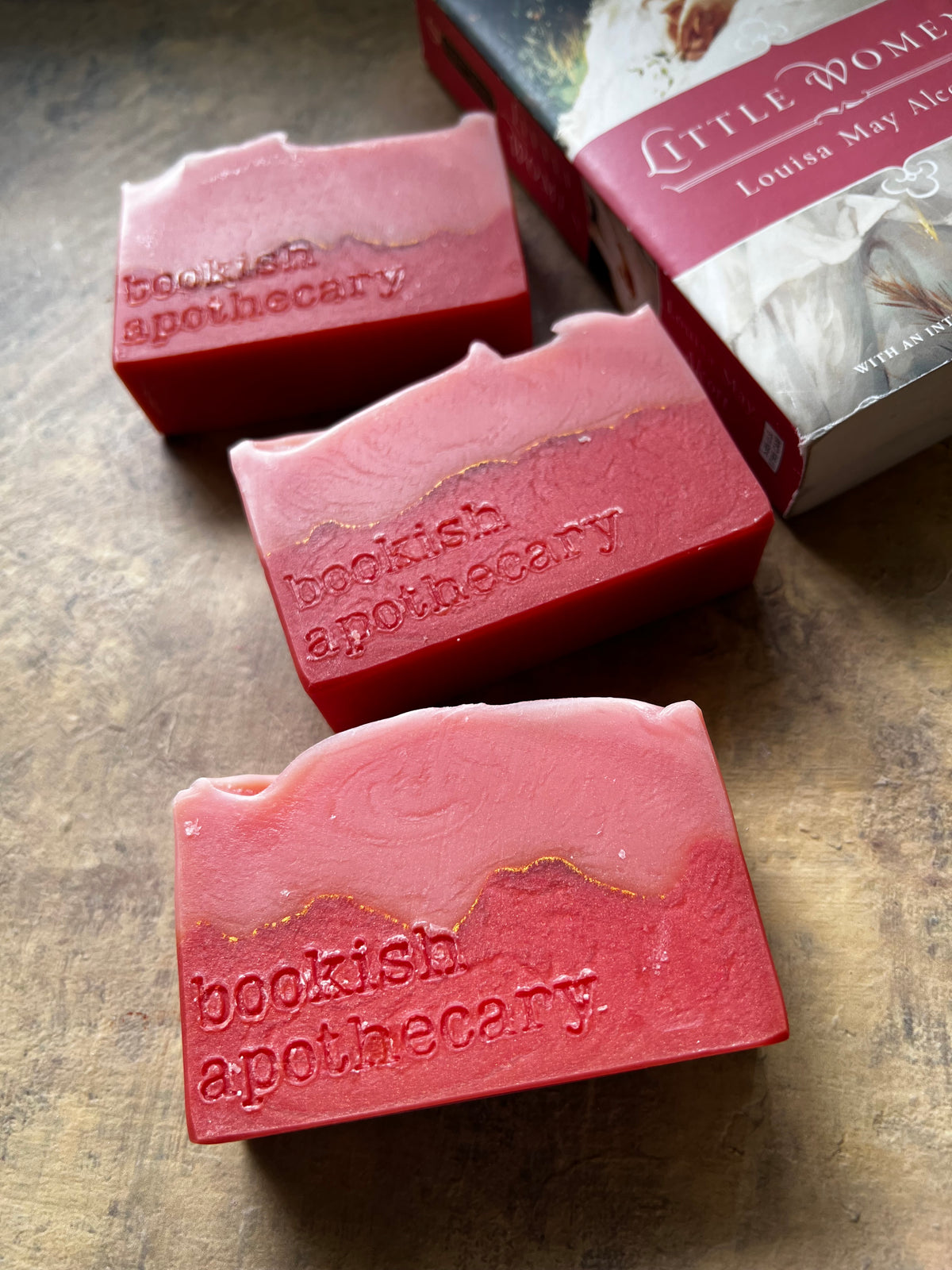 Three pink soap bars embossed with 'bookish apothecary' are arranged on a textured brown surface next to a book titled 'Little Women' by Louisa May Alcott. The soap has a swirled pink and red pattern with a thin gold line running through it.
