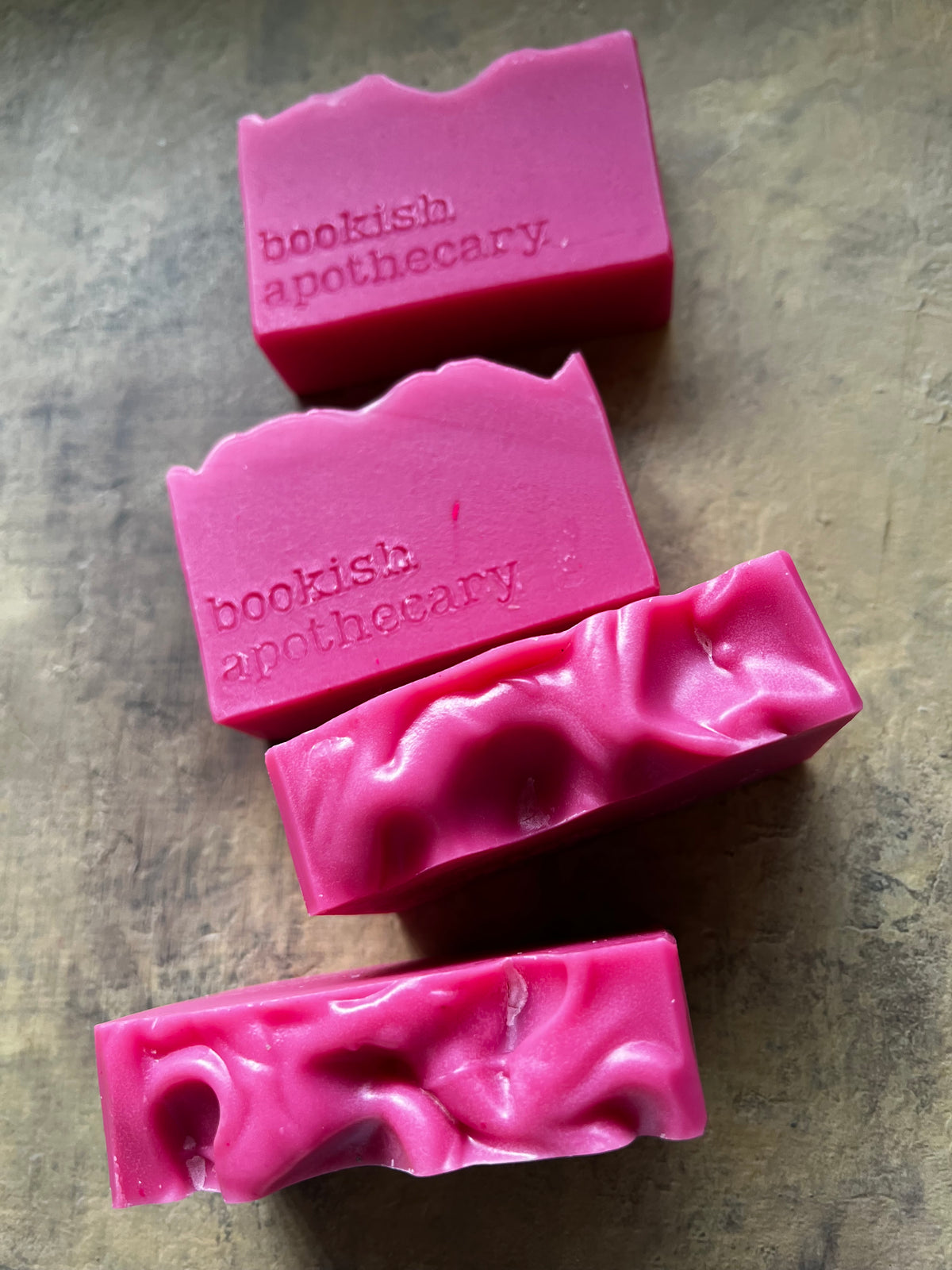 Four pink handmade soap bars with a wavy, textured top edge, embossed with the brand name 'bookish apothecary', arranged diagonally on a textured gray surface.