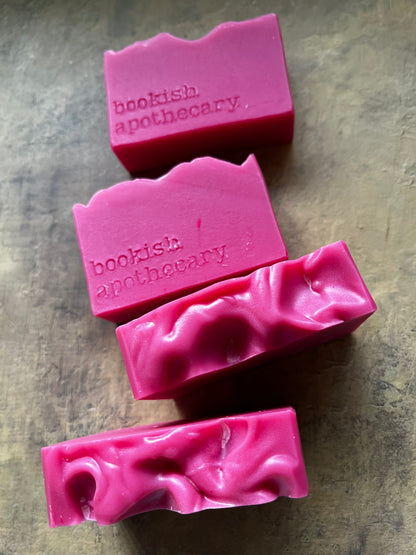 Four pink handmade soap bars with a wavy, textured top edge, embossed with the brand name 'bookish apothecary', arranged diagonally on a textured gray surface.