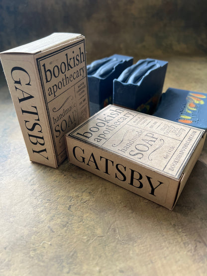 Two rectangular boxes of Gatsby Bookish Apothecary soap stacked on a textured concrete surface, with a stack of blue soap bars in the background.