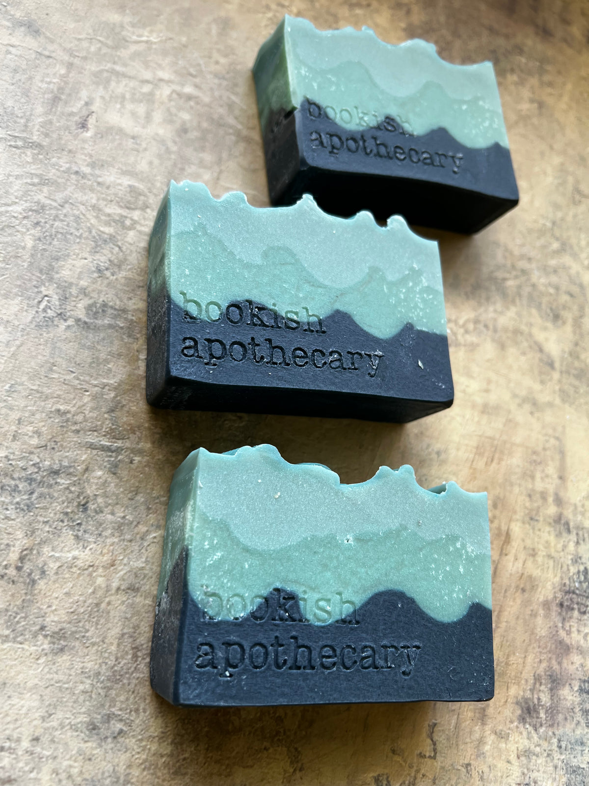 Three rectangular artisan soap bars with a wavy, layered design in mint green and dark charcoal gray, each embossed with the brand name 'bookish apothecary', arranged diagonally on a textured brown wooden surface.