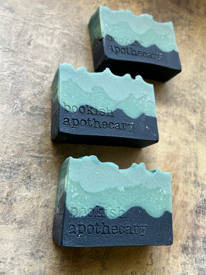 Three rectangular artisan soap bars with a wavy, layered design in mint green and dark charcoal gray, each embossed with the brand name 'bookish apothecary', arranged diagonally on a textured brown wooden surface.