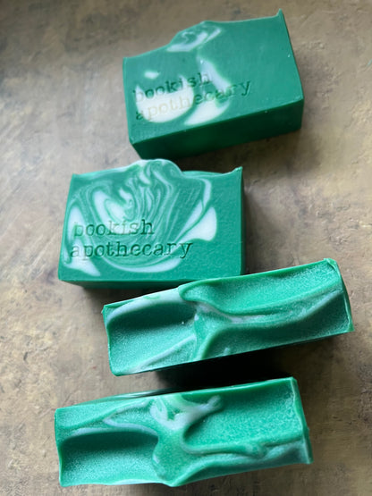 Four green soap bars with white marbled swirls, featuring the text 'bookish apothecary' stamped on two of the bars, arranged on a textured brown surface.
