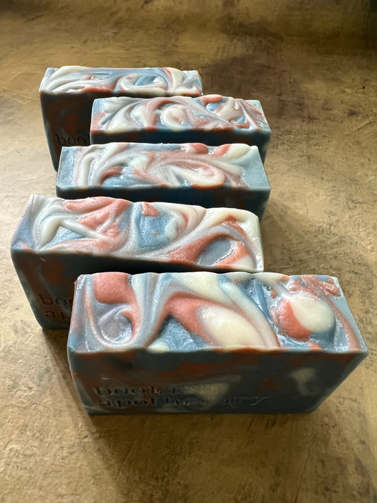 Five handmade marbled soap bars with swirls of gray, white, and terracotta orange, arranged diagonally on a textured brown surface.