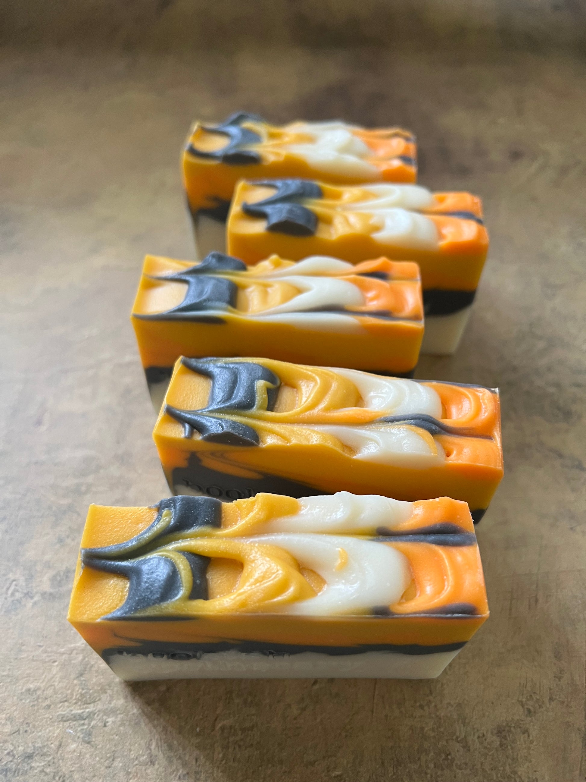 Five rectangular handmade soap bars with a marbled swirl pattern of orange, white, and dark gray, displayed on a textured beige surface.