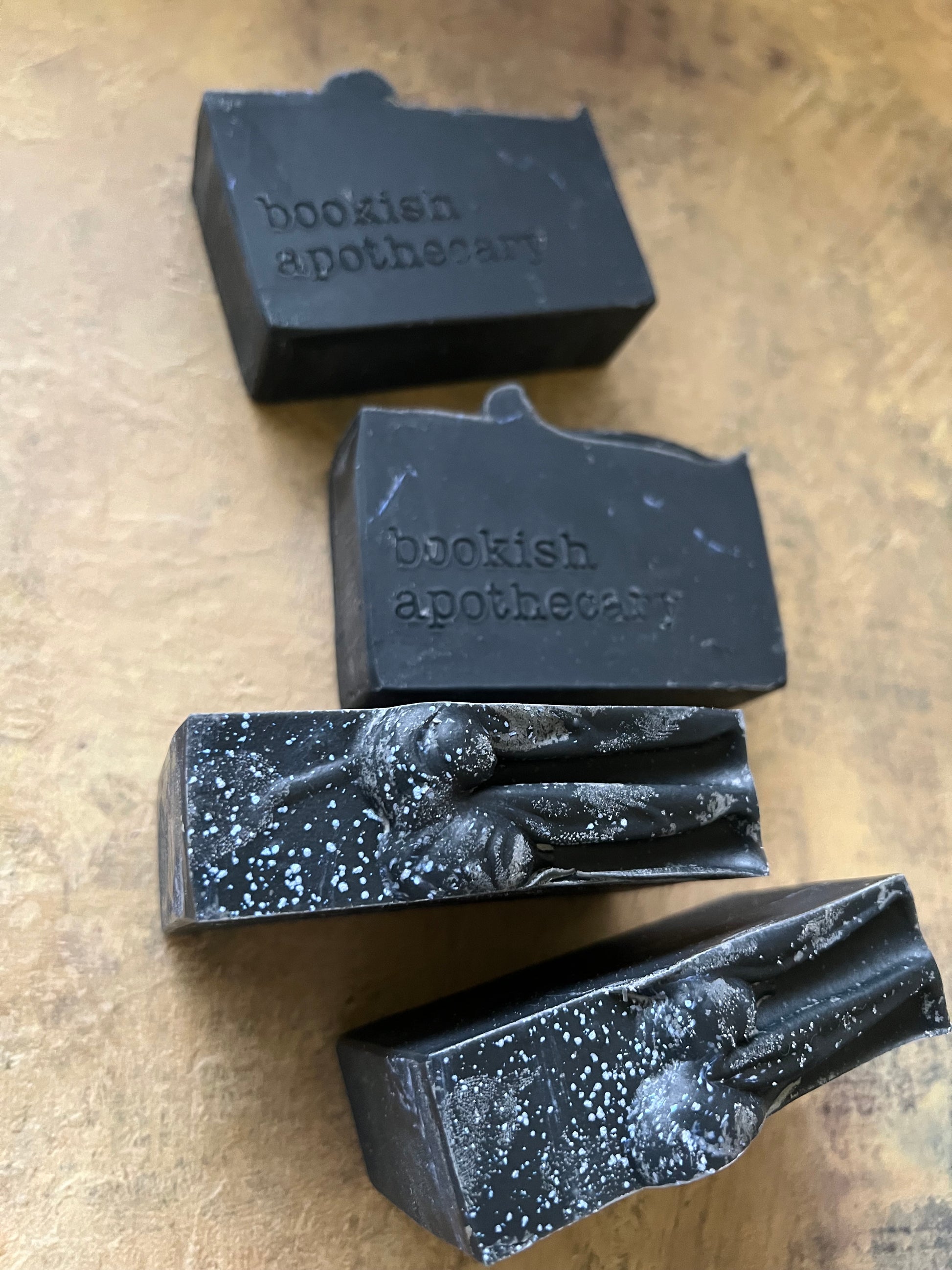 Four dark, rectangular handmade soap bars with white speckles and the brand name 'bookish apothecary' embossed on them, arranged on a textured tan surface.