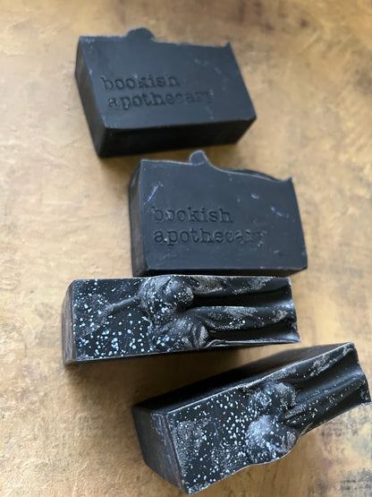 Four dark, rectangular handmade soap bars with white speckles and the brand name 'bookish apothecary' embossed on them, arranged on a textured tan surface.