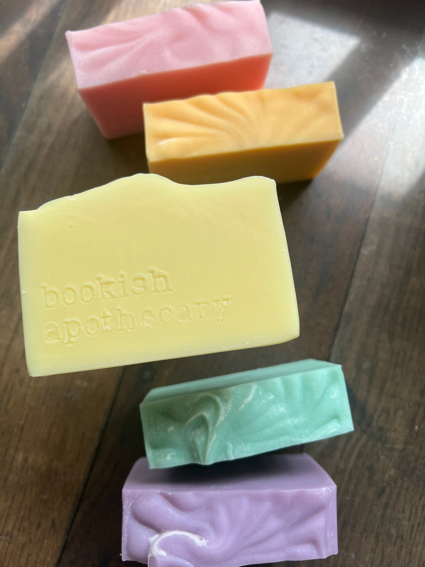 Five colorful soap bars on a wooden surface, with one bar labeled 'Bookish Soap Company'.