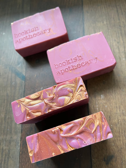 Four pink soap bars with gold marbling on a wooden surface, branded 'bookish apothecary'.
