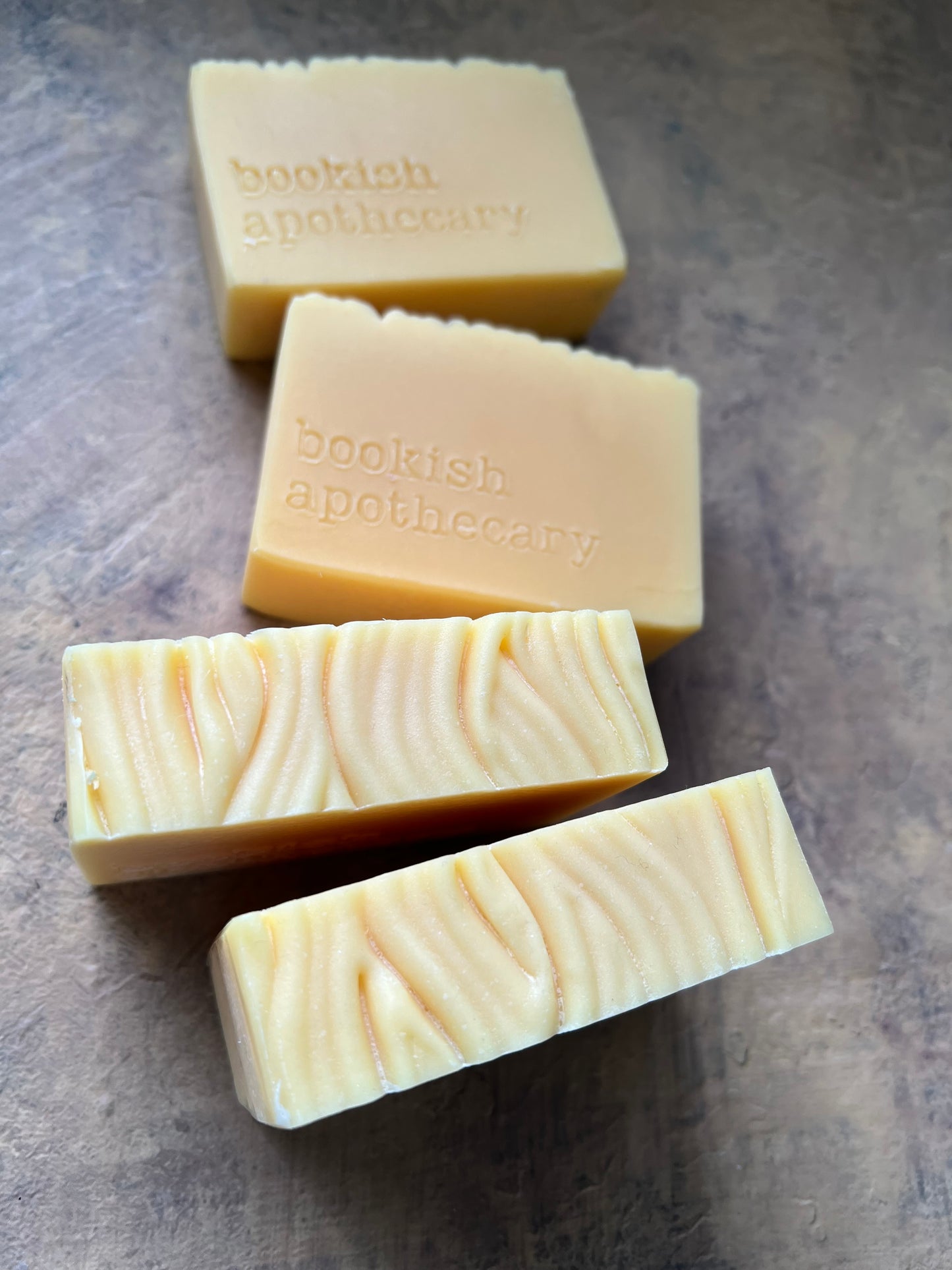 Four handmade soap bars in pale creamy yellow with textured and wavy cuts, stamped with the brand name 'bookish apothecary', arranged on a textured gray surface.