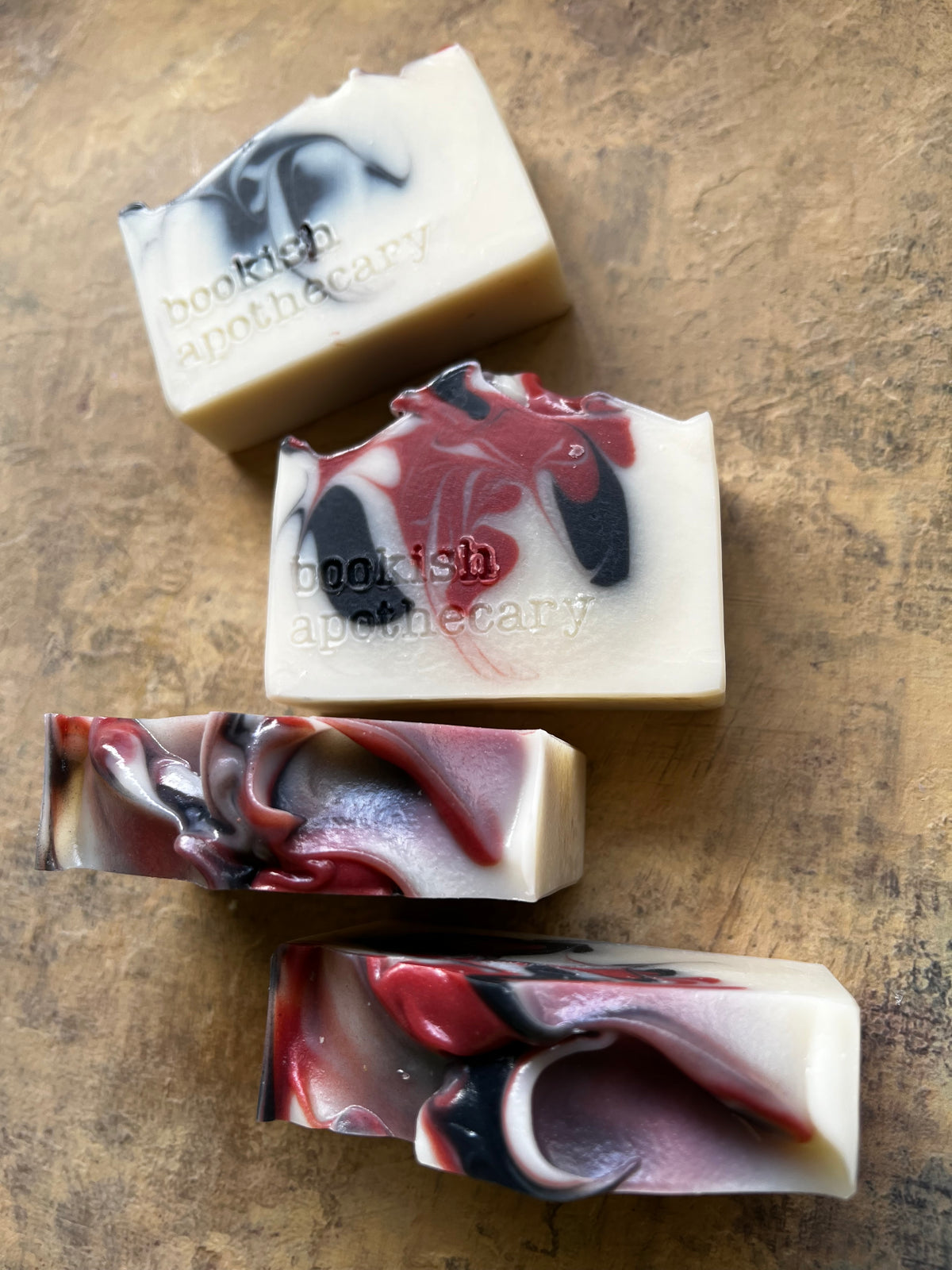 Four artisanal soap bars with marbled swirls of cream, red, black, and gray, arranged diagonally on a textured brown surface. Two bars feature the brand name 'bookish apothecary' embossed on them.