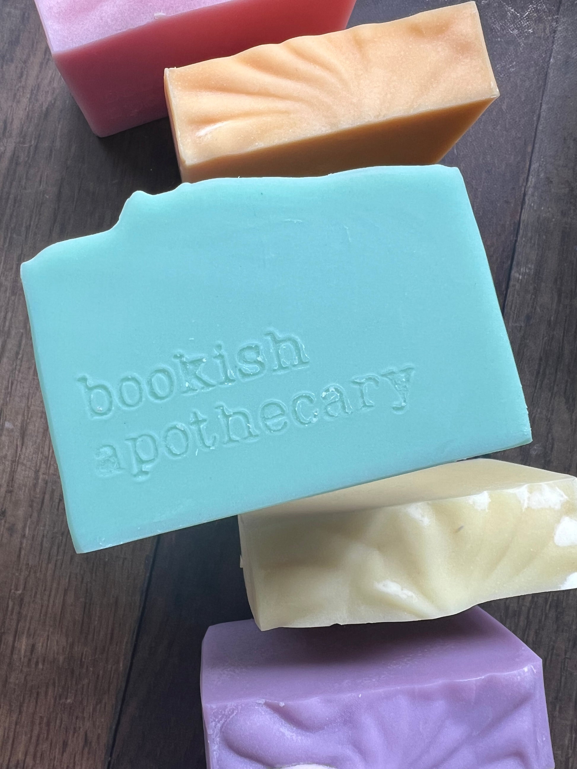 Five bars of soap in different colors with 'bookish apothecary' branding on a wooden surface.