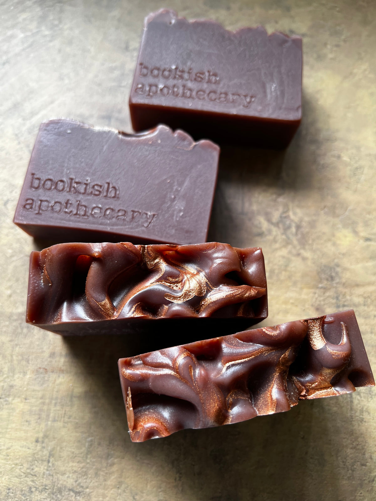 Four bars of handmade soap from Bookish Apothecary arranged on a textured metallic surface. Two bars are shown upright with the brand name embossed on top, and two are shown in profile to reveal their swirled, layered texture in dark brown and coppery metallic tones.