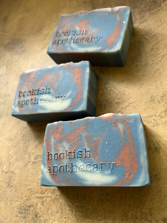 Three rectangular bars of soap with a blue-gray base and orange swirls, printed with the brand name 'bookish apothecary', displayed on a textured beige surface.