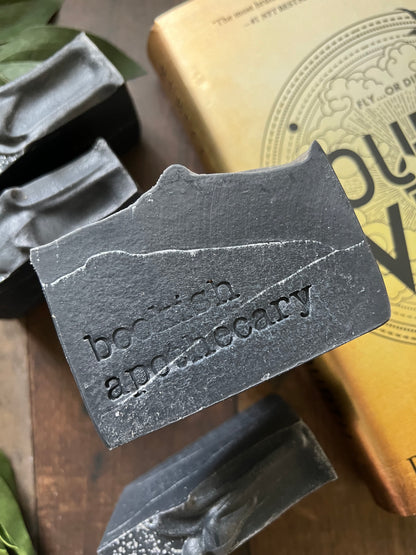 A bar of handmade soap with a gray color and speckled with dark dots, placed on a wooden surface with green leaves in the background and a label that reads 'COLLECTOR EDITION online exclusive'.