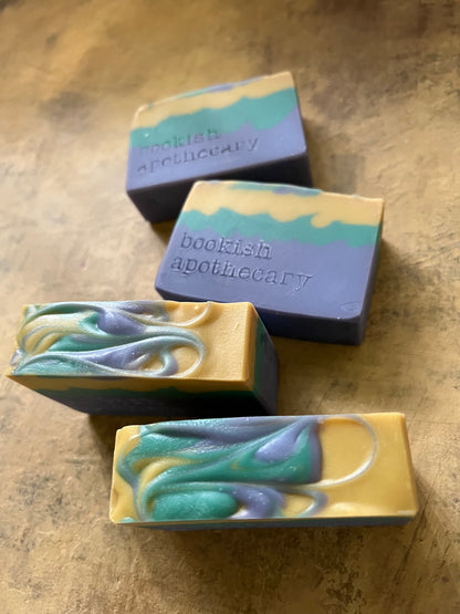 Four handmade artisanal soaps with purple, teal, and yellow marbled swirls, arranged on a textured brown surface. Two soaps feature the brand name 'bookish apothecary' stamped on the top.
